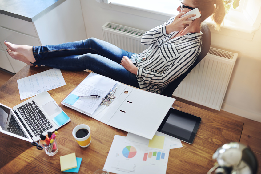 Relaxed confident female entrepreneur reclining back in her chair with her bare feet on the desk talking to a client on her mobile phone, high angle view on desk with paperwork, charts and laptop